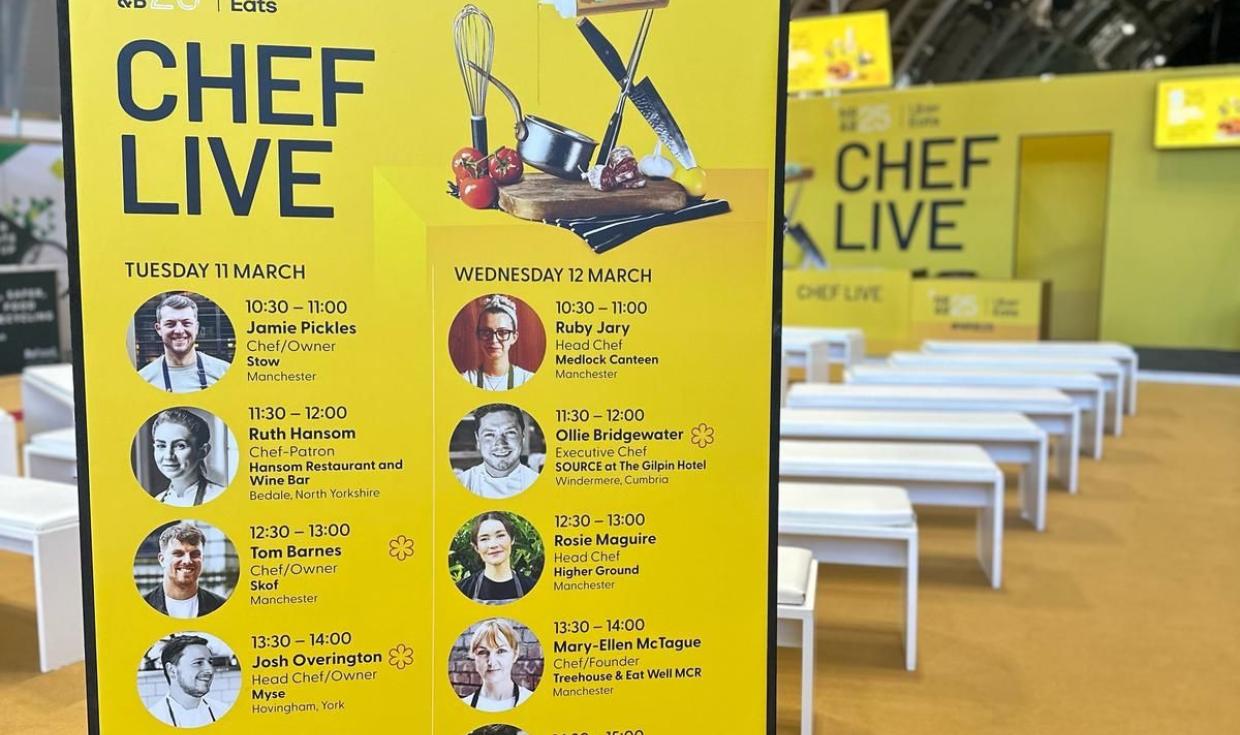 Information board, displaying line up information for Chef Live stage against a yellow background. Chef Live stage and seating in background.