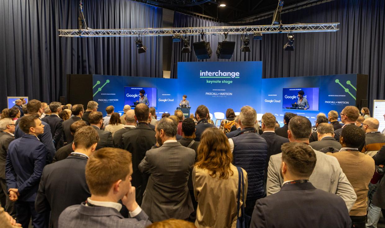 Keynote Speakers on stage on corner of Exchange Hall with blue screens as backdrop, with a crowd watching.