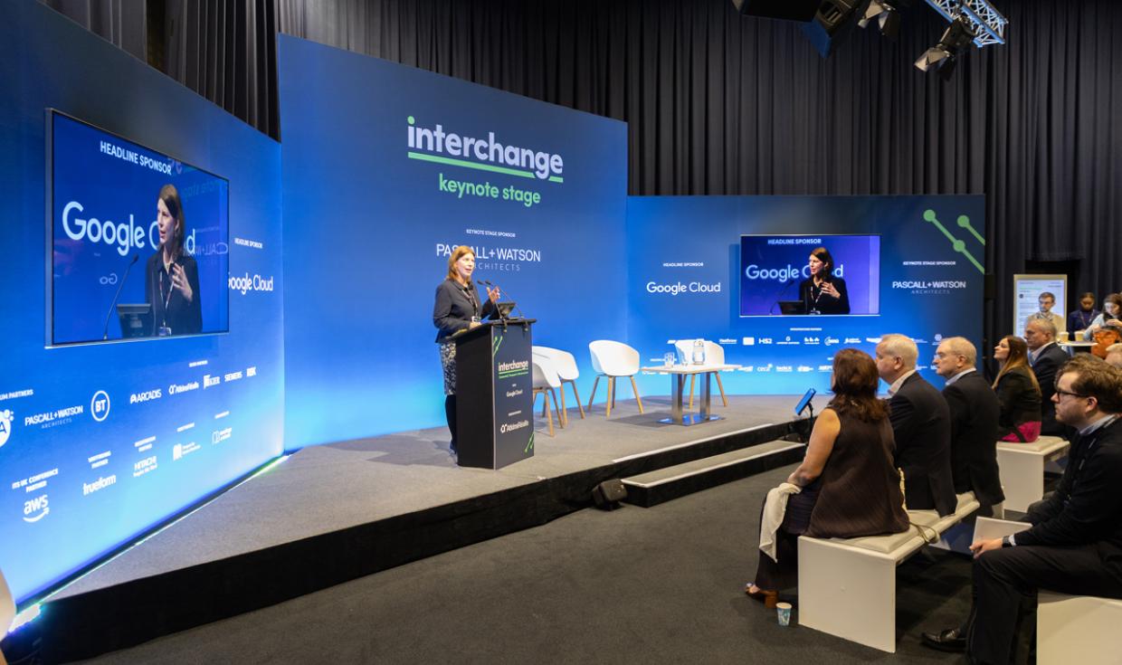 Keynote Speakers on stage on corner of Exchange Hall with blue screens as backdrop, with a delegates sat watching.