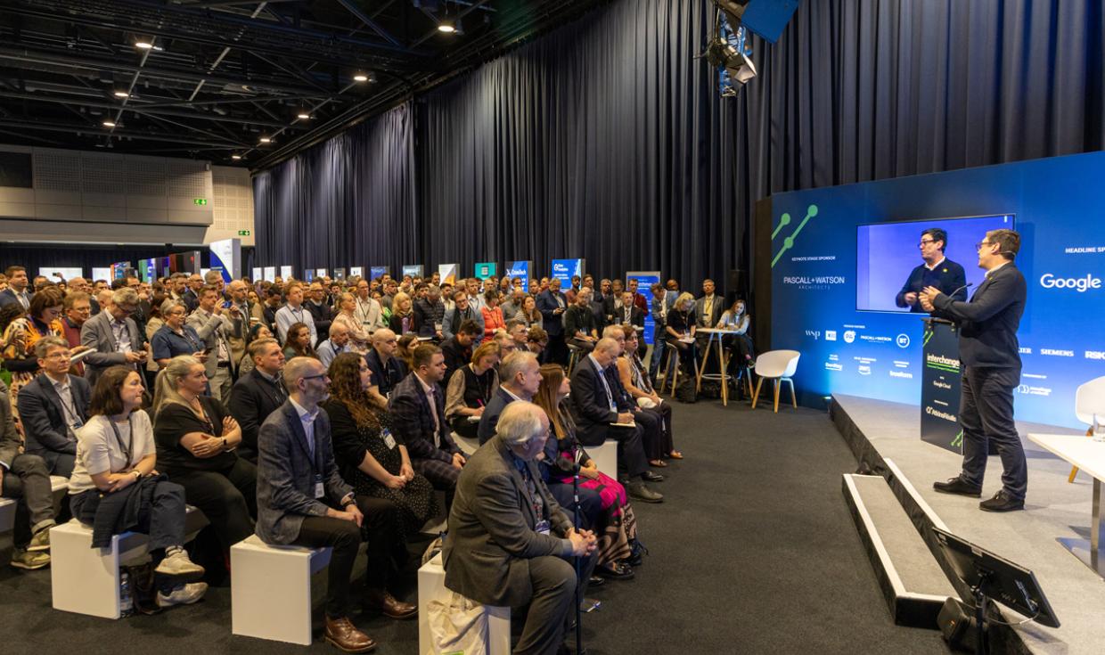 Keynote Speakers on stage on corner of Exchange Hall with blue screens as backdrop, with a crowd sat on benches watching.