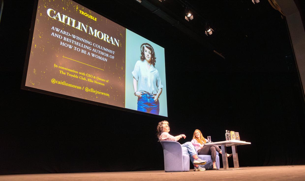 An angled shot showing Caitlin Moran and host sat on stage, with a large format screen behind them that reads 'Caitlin Moran'